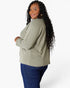 A woman with long black curly hair is shown from the back and side, wearing a light sage green long-sleeve top and dark blue jeans against a plain white studio background.