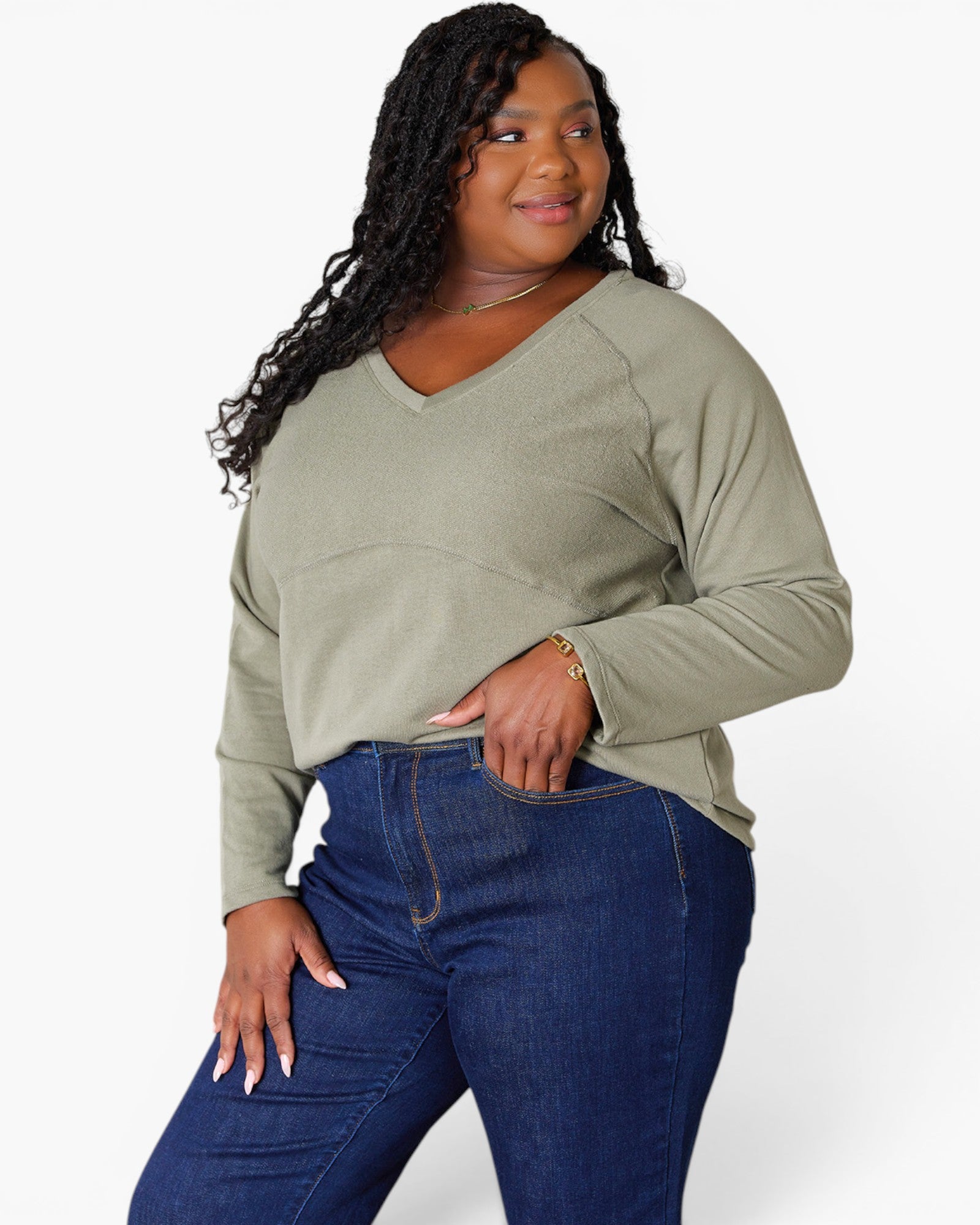 A woman modeling a sage green long-sleeve V-neck top with dark blue jeans against a plain white studio background.