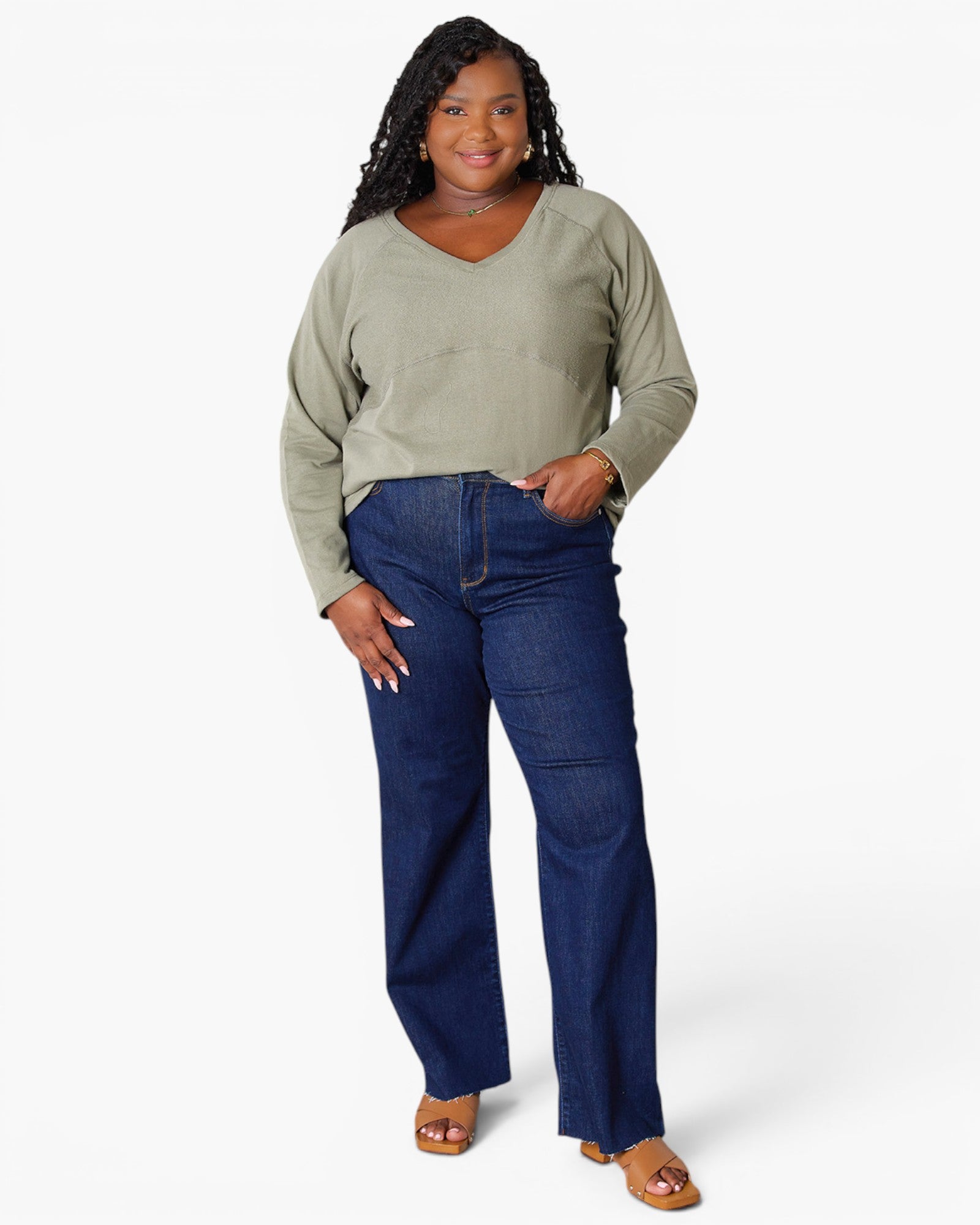 A woman modeling a sage green long-sleeve V-neck top and dark blue wide-leg jeans with brown sandals, standing against a light gray studio background.