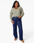 A woman modeling a sage green long-sleeve V-neck top and dark blue wide-leg jeans with brown sandals, standing against a light gray studio background.