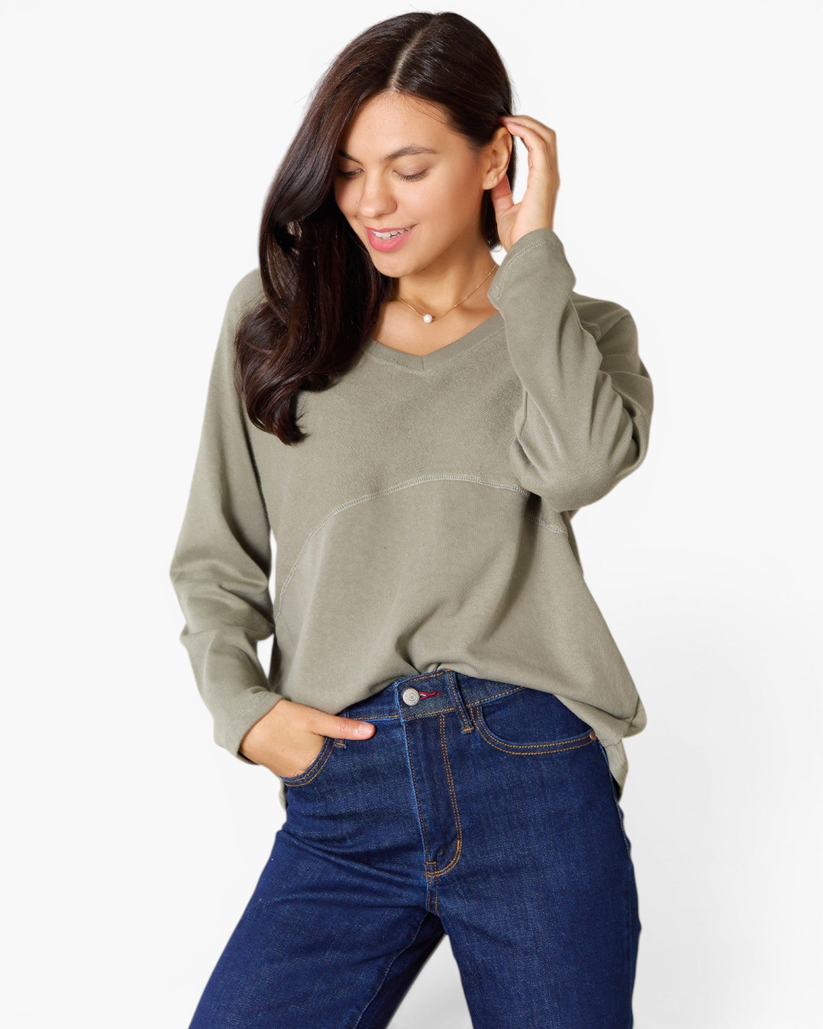 A woman wearing a sage green long-sleeve V-neck top and dark blue jeans, posing against a light gray studio background.