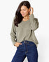 A woman wearing a sage green long-sleeve V-neck top and dark blue jeans, posing against a light gray studio background.