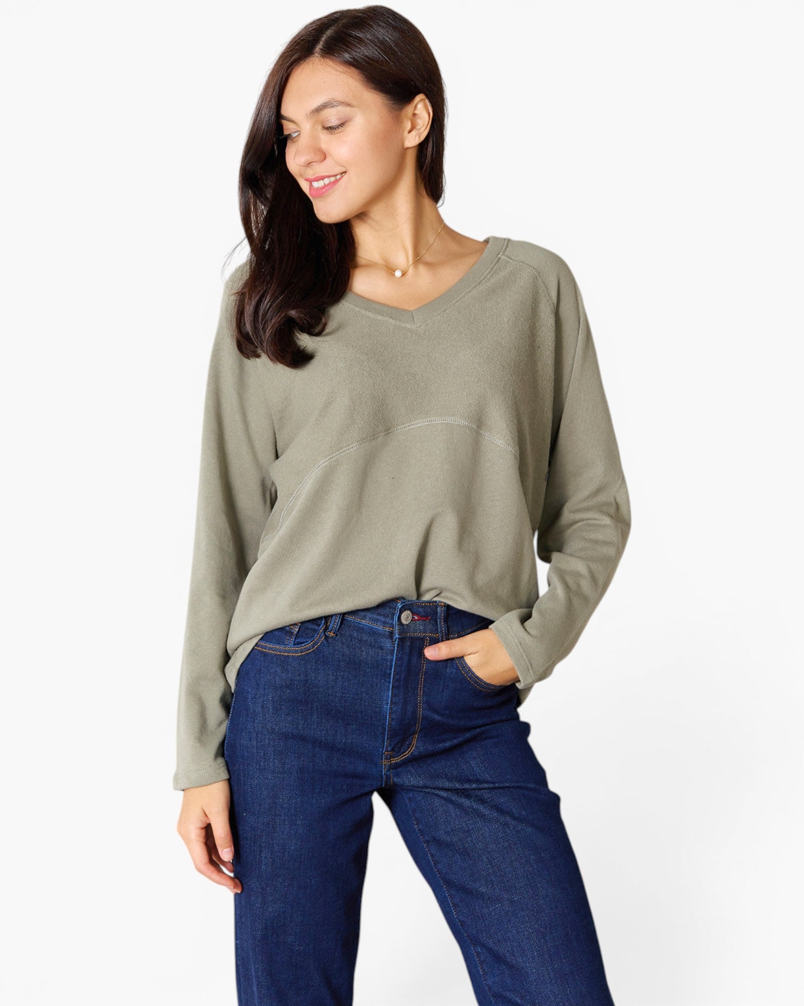 A woman wearing a sage green long-sleeve knit top and dark blue jeans, standing against a plain white studio background.