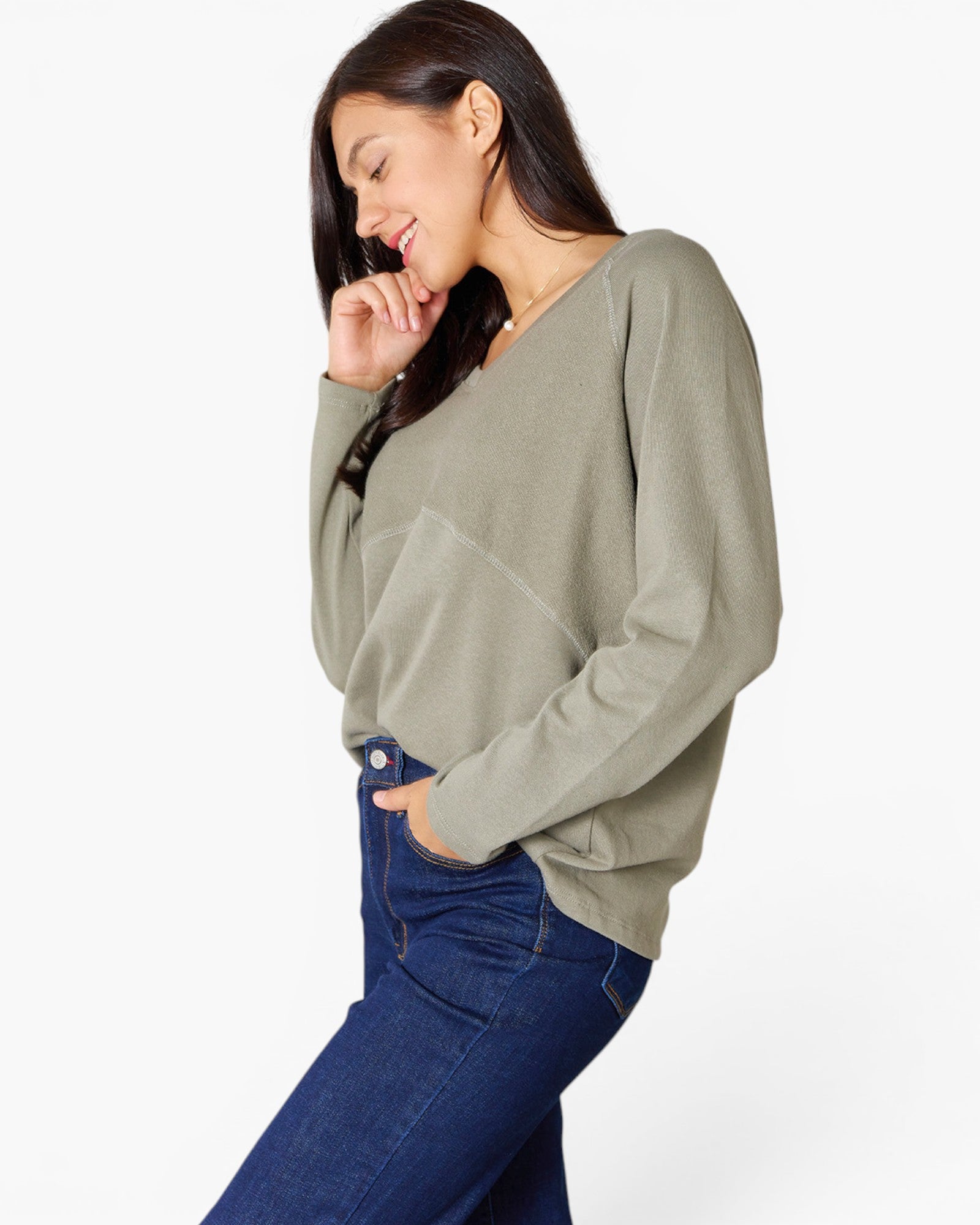 A woman wearing a sage green long-sleeve knit top and dark blue jeans, posing against a light gray studio background.