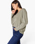 A woman wearing a sage green long-sleeve knit top and dark blue jeans, posing against a light gray studio background.
