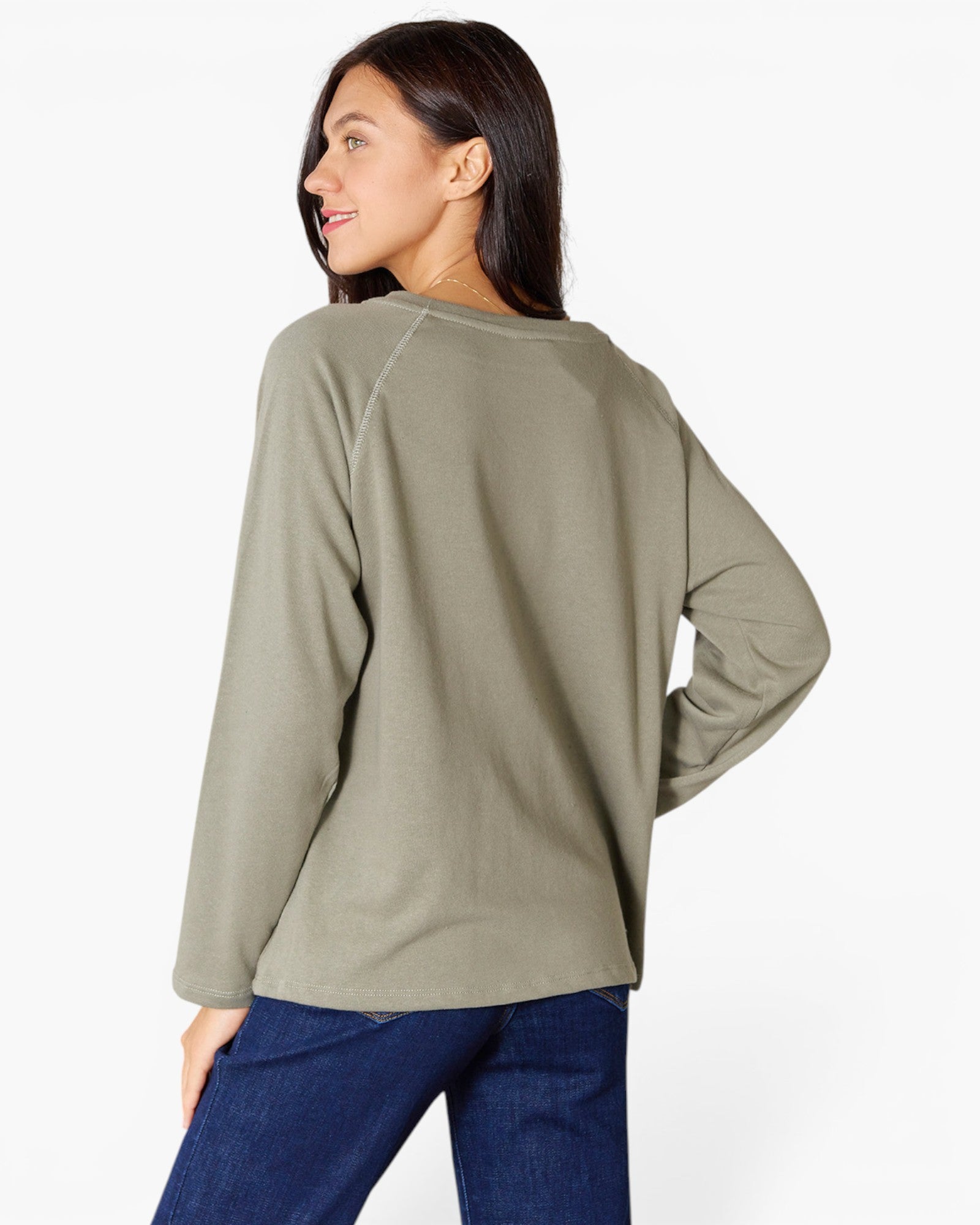 A woman shown from the back wearing a sage green long-sleeve raglan top and dark blue jeans, photographed against a clean white studio background.