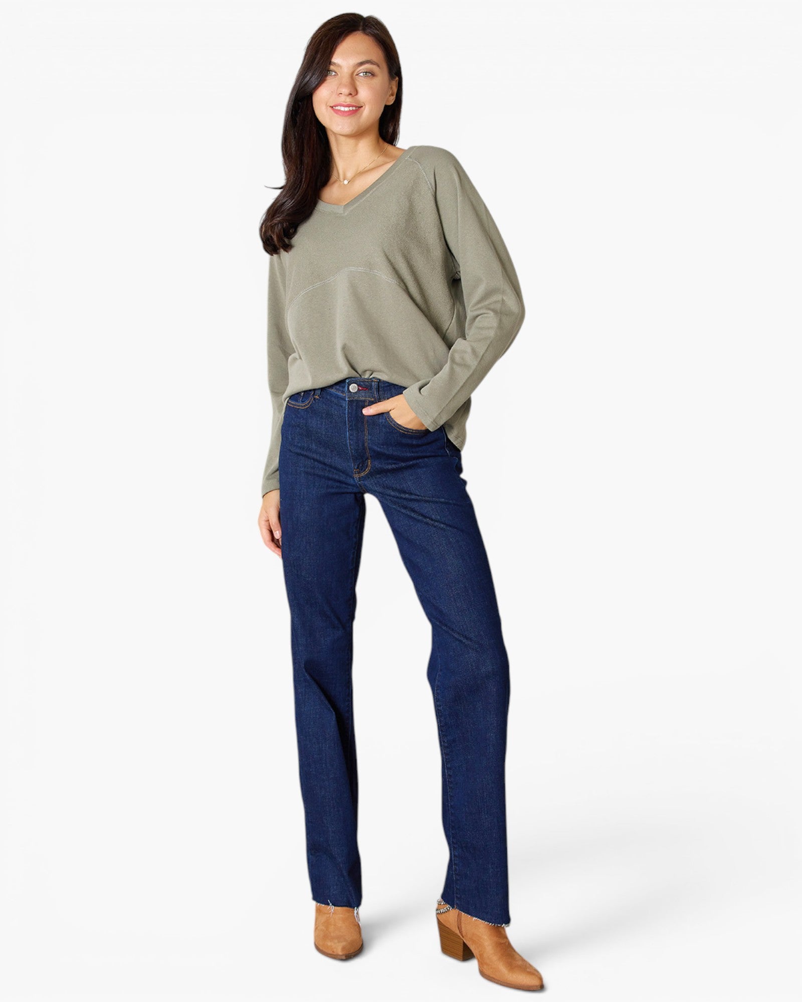 A woman modeling a sage green long-sleeve V-neck top and dark blue straight-leg jeans with tan ankle boots, standing against a light gray studio background.