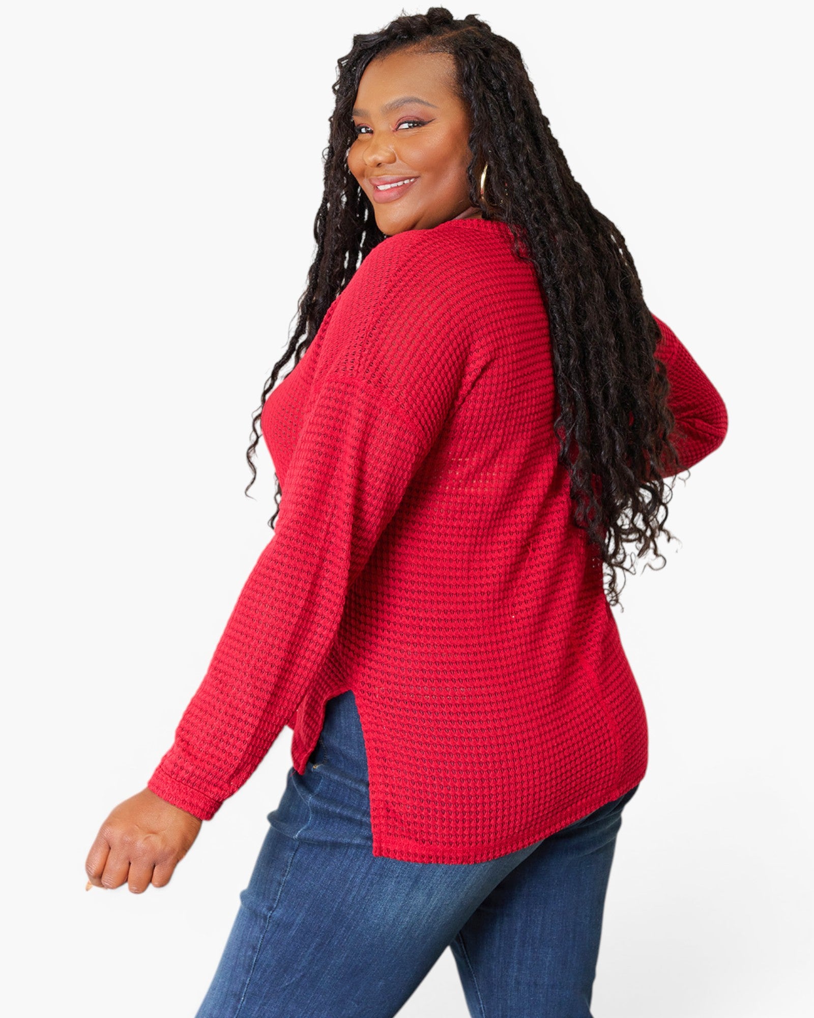 A woman with long black curly hair is shown from a three-quarters back view, wearing a bright red waffle-knit long-sleeve top and blue jeans against a plain white studio background.