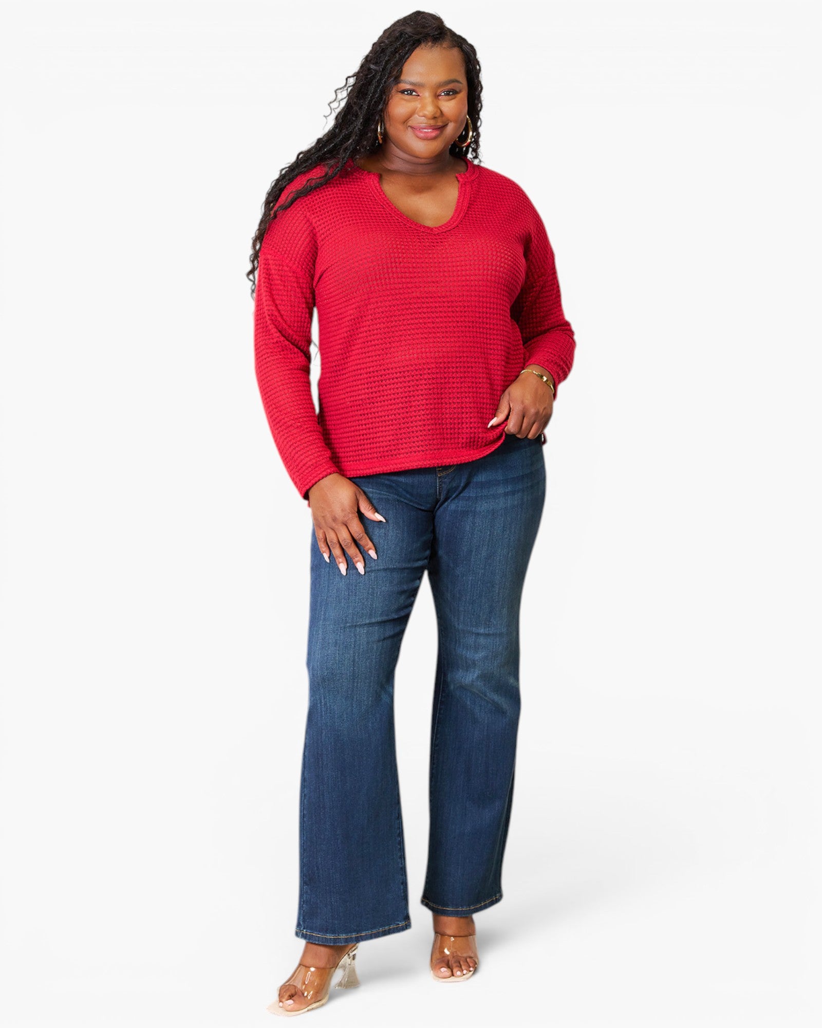 A woman modeling a red ribbed long-sleeve V-neck top with dark blue flare jeans and clear high-heeled sandals, standing against a light gray studio background.