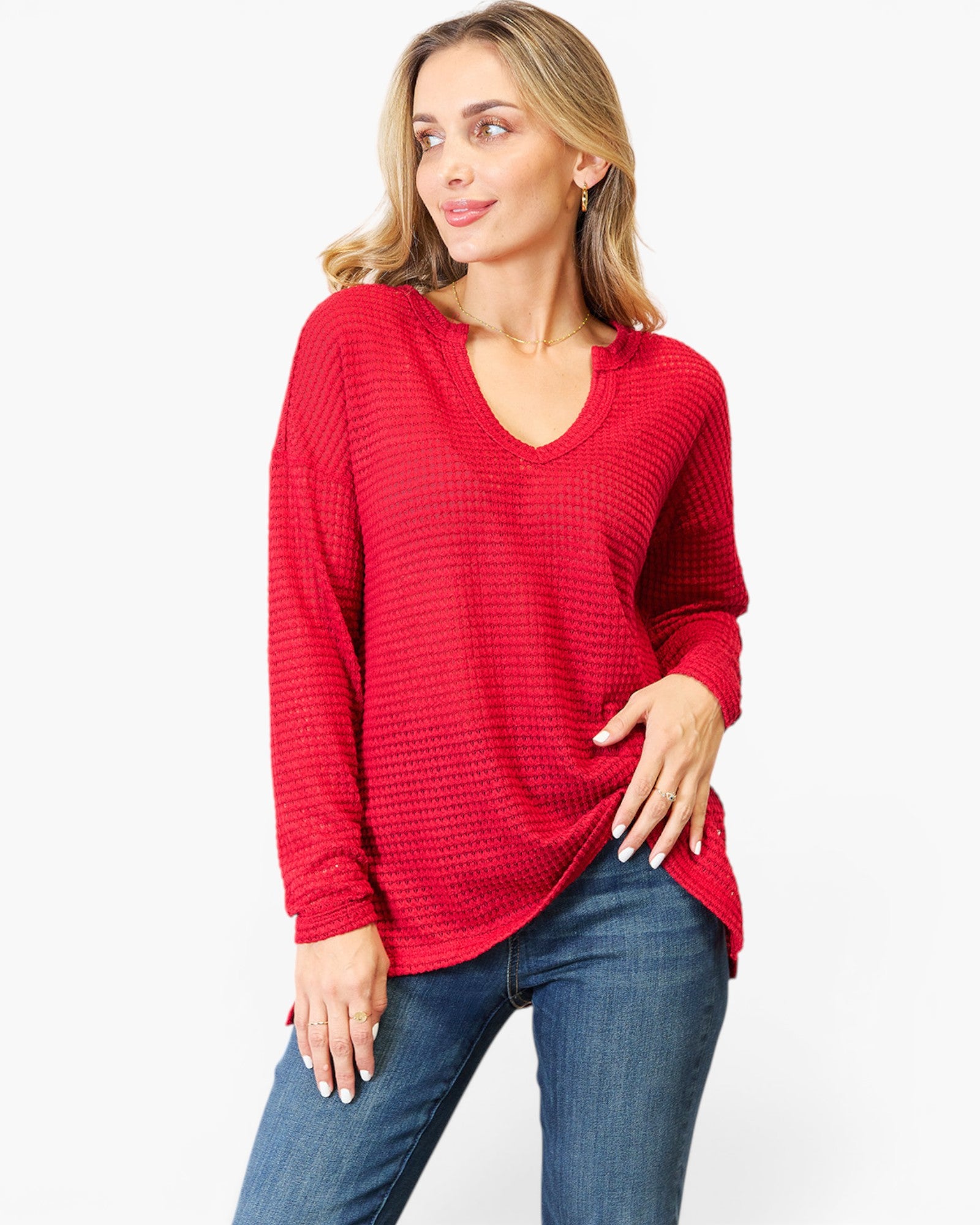 A woman modeling a red waffle-knit long-sleeve V-neck top with blue jeans against a plain white background.