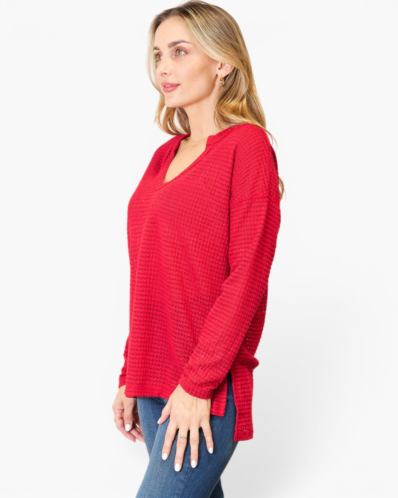 A woman wearing a red waffle-knit long-sleeve top with a split V-neckline, paired with blue jeans, photographed against a plain white studio background.