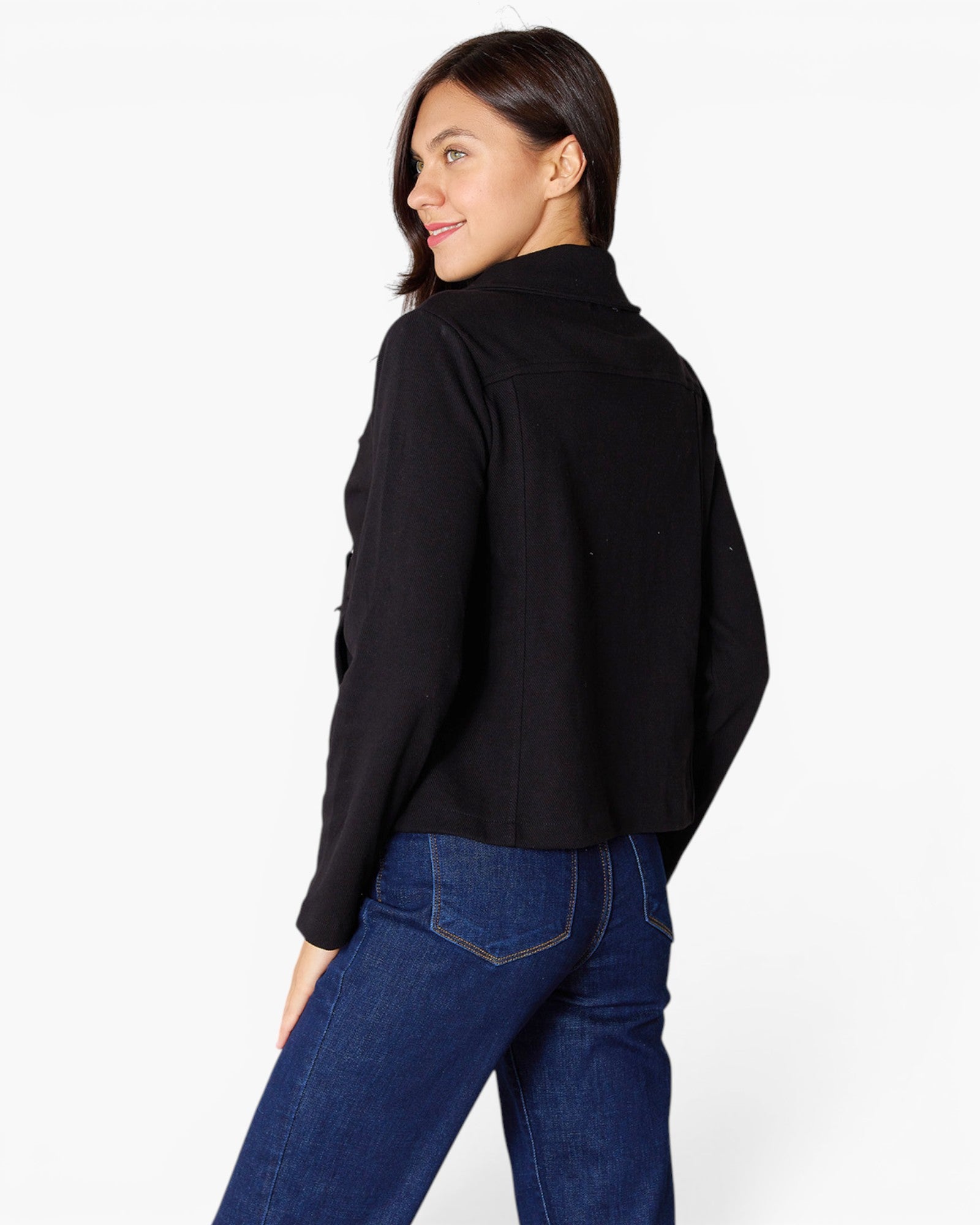 A woman wearing a black cropped long-sleeve shirt and blue jeans, photographed from the back against a plain white studio background.