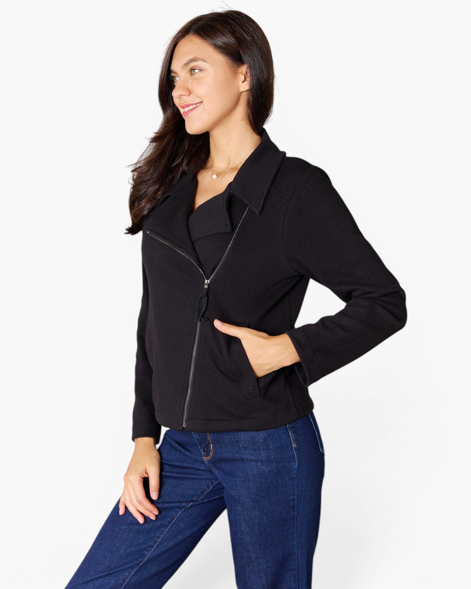A woman wearing a black zip-front jacket with a collar and blue jeans, standing against a light gray studio background.