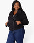 A woman modeling a black zip-up jacket with a wide collar and front pockets, paired with blue jeans, against a plain light gray studio background.