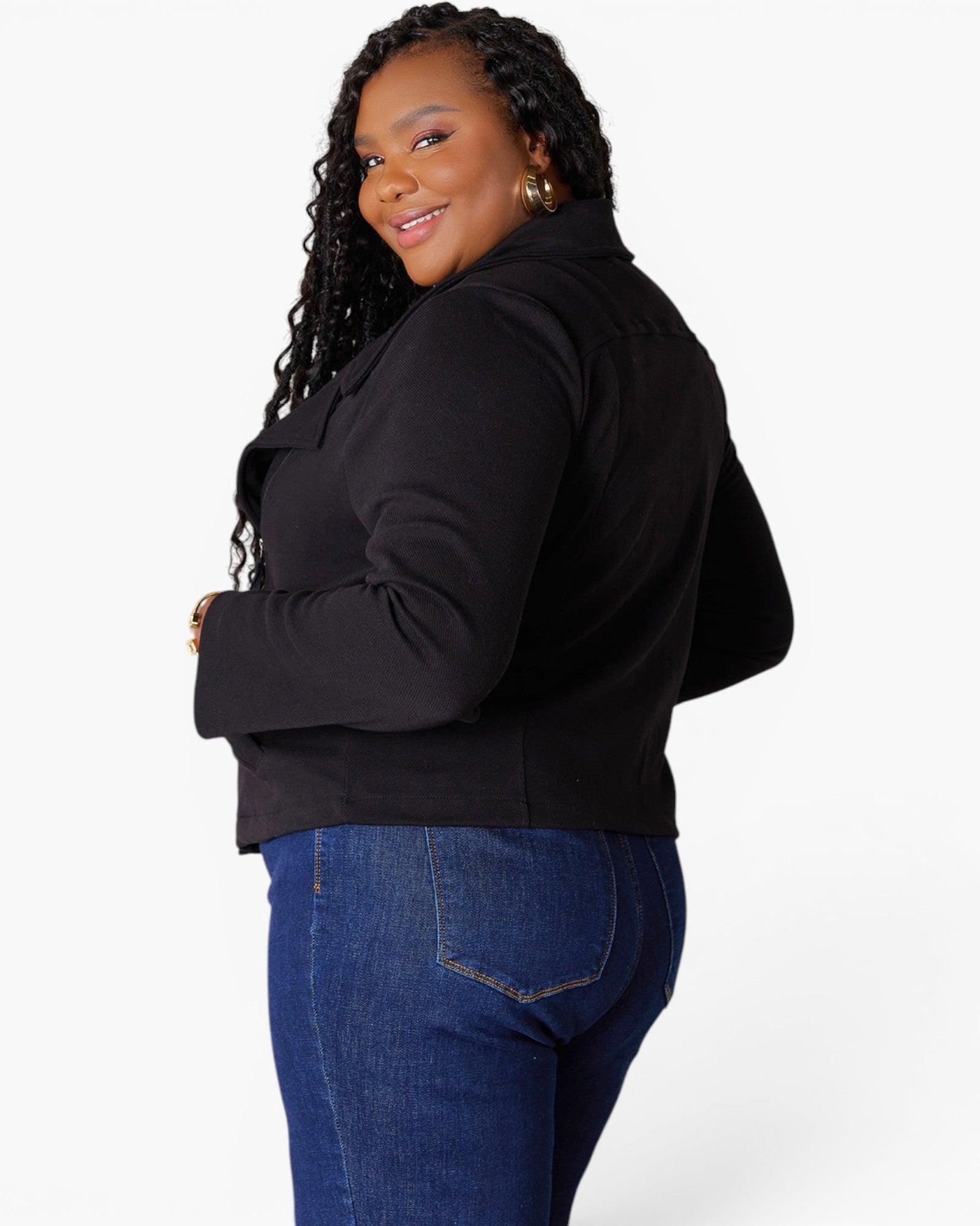 A woman modeling a black long-sleeve cropped jacket with a high collar, paired with blue jeans, shown from a three-quarters back view against a light gray studio background.