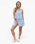 A woman modeling a light blue and white striped two-piece set consisting of a sleeveless peplum-style top with spaghetti straps and matching shorts, paired with white slide sandals, against a plain white studio background.