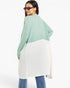 A woman wearing a color-blocked long-sleeve tunic top with a sage green upper section and an off-white lower section, paired with blue jeans against a plain white studio background.