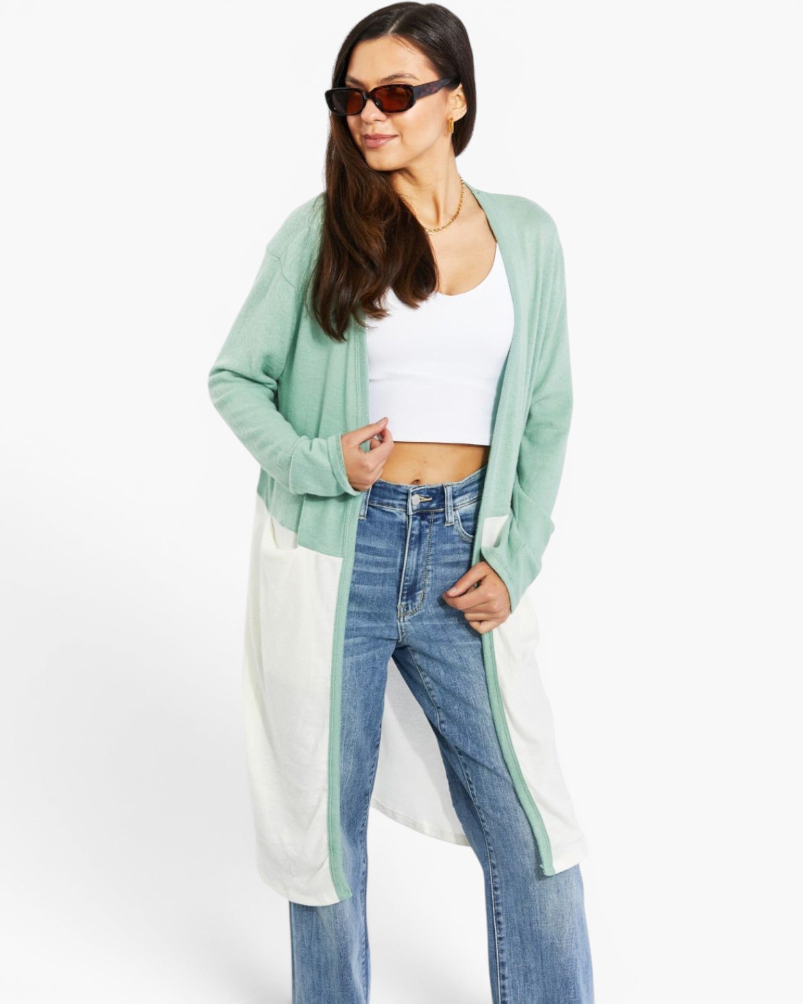A woman modeling a long, color-blocked cardigan with sage green, white, and light blue panels, worn over a white crop top and blue jeans against a plain white background.