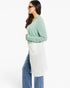 A woman wearing a sage green long-sleeve cardigan over a white tunic top, paired with blue jeans, shown from a side profile against a plain white background.