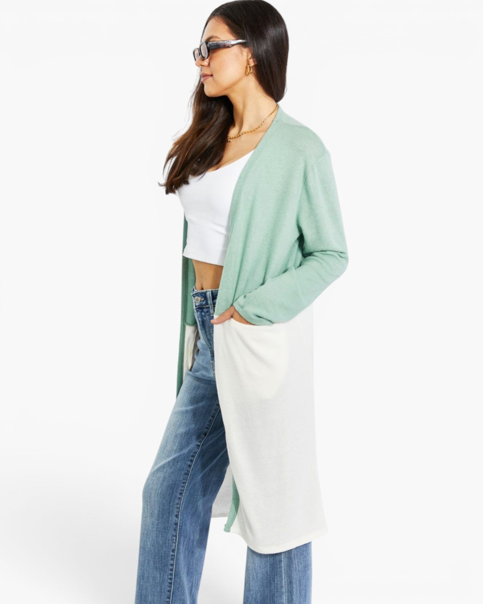 A woman wearing a long, color-blocked cardigan with a sage green upper section and a white lower section, styled with a white crop top and blue jeans against a plain white background.