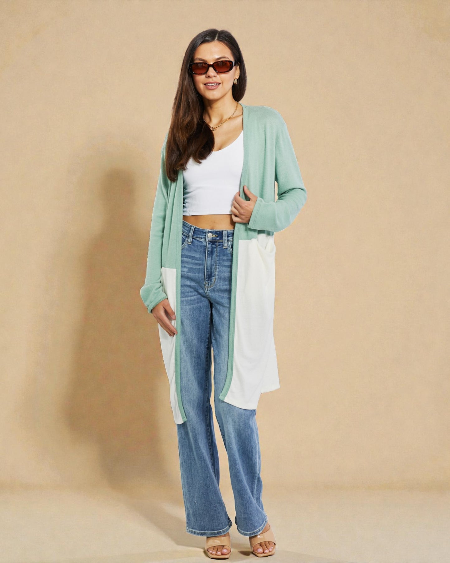 Woman wearing a green cardigan, white top, blue jeans, and sunglasses on a beige background