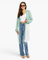 A woman modeling a long, color-blocked cardigan with sage green, white, and light blue panels, worn over a white crop top and blue jeans, on a plain white studio background.