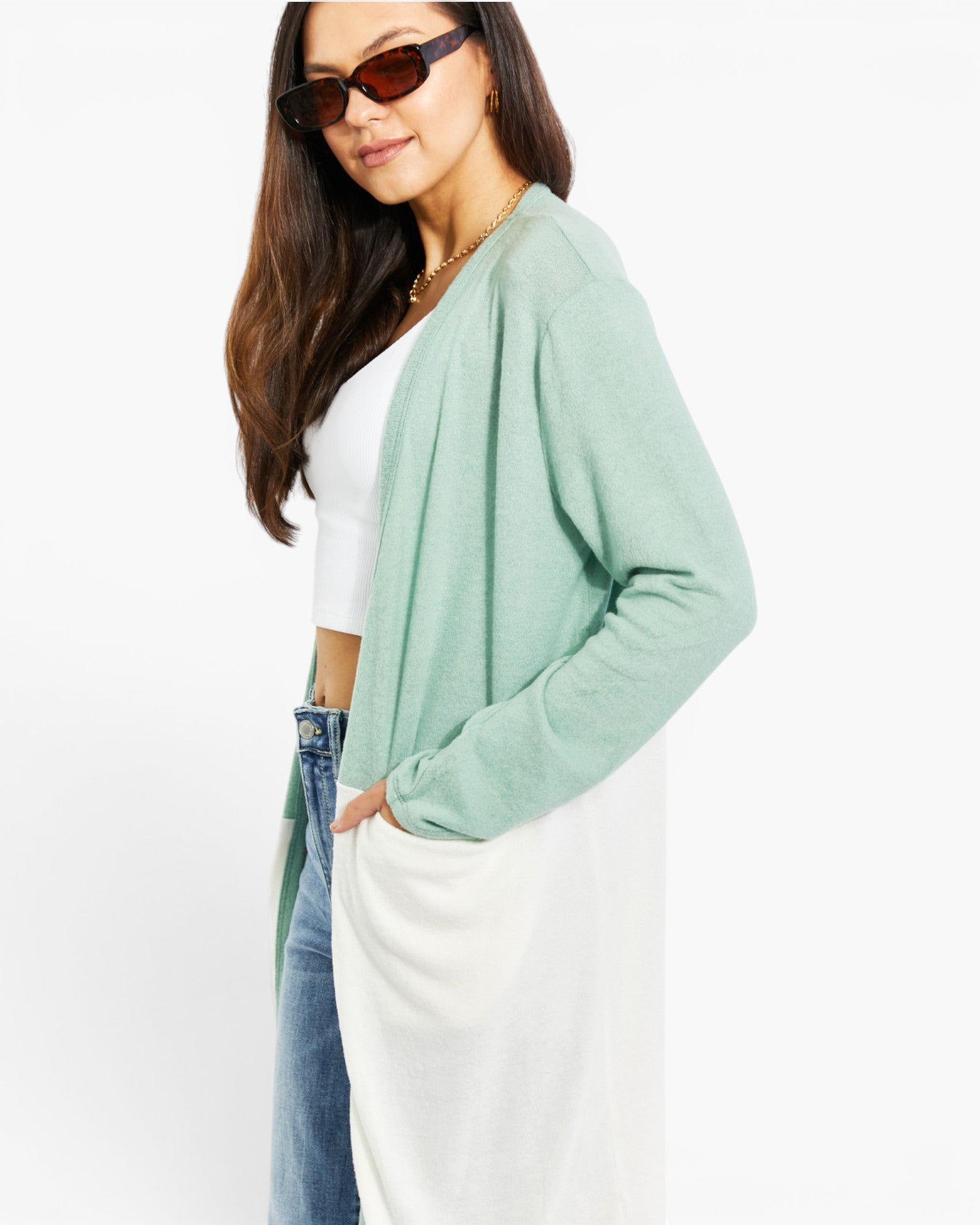 A woman wearing a light sage green open-front cardigan over a white top and blue jeans, photographed against a plain white studio background.