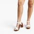 White platform sandals with ankle straps and brown block heels, shown on a model's feet against a light gray studio background.