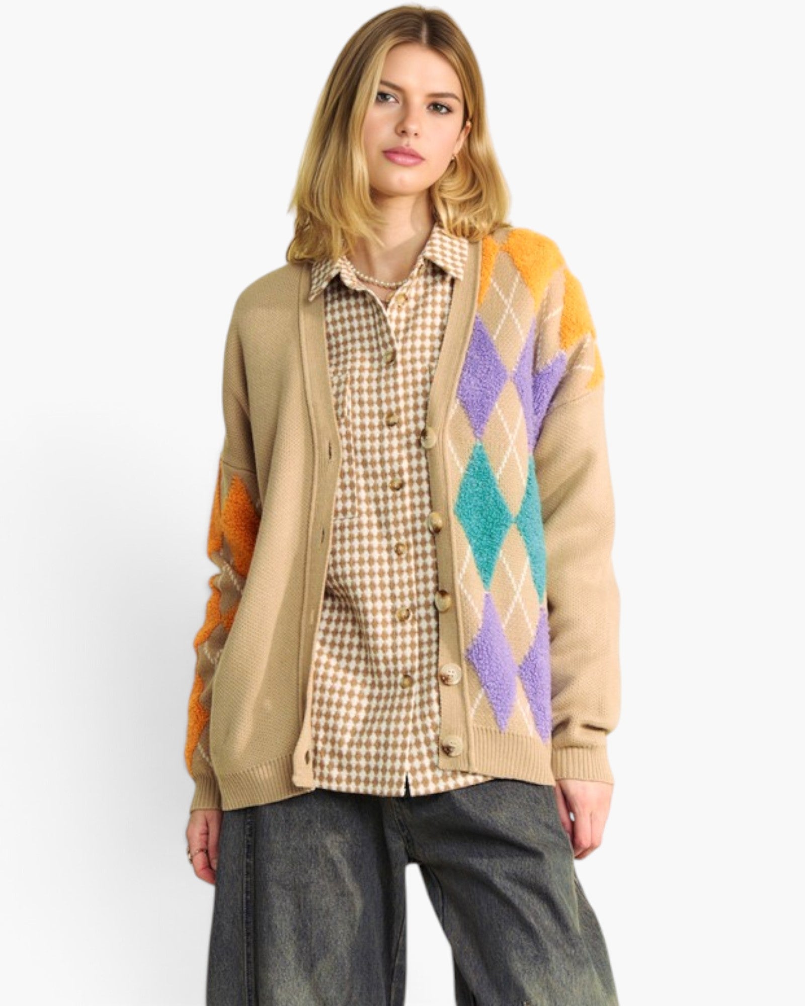 A woman modeling a beige cardigan with a colorful argyle pattern on the front and sleeves, worn over a brown and beige checkered shirt and dark gray jeans, against a plain white background.