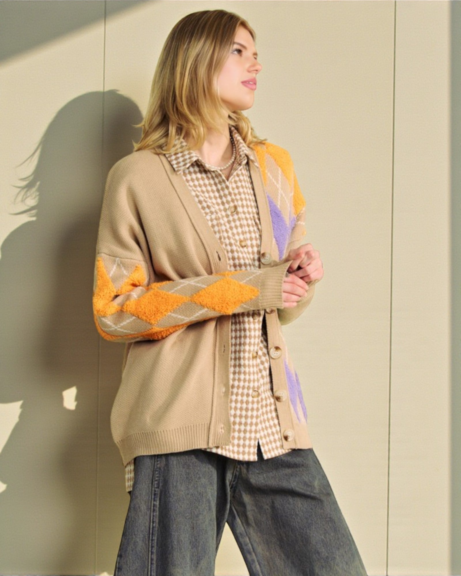 Person wearing a beige cardigan with orange and gray pattern, plaid shirt, and jeans against a neutral wall.
