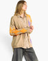 A woman wearing a beige cardigan with orange and purple argyle knit patterns over a brown and white checkered shirt, paired with dark gray jeans, against a plain white background.