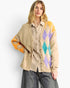 A woman models a beige button-front cardigan with colorful argyle and floral knit patterns on the sleeves and side, worn over a beige-and-white check shirt and gray jeans against a light gray studio background.