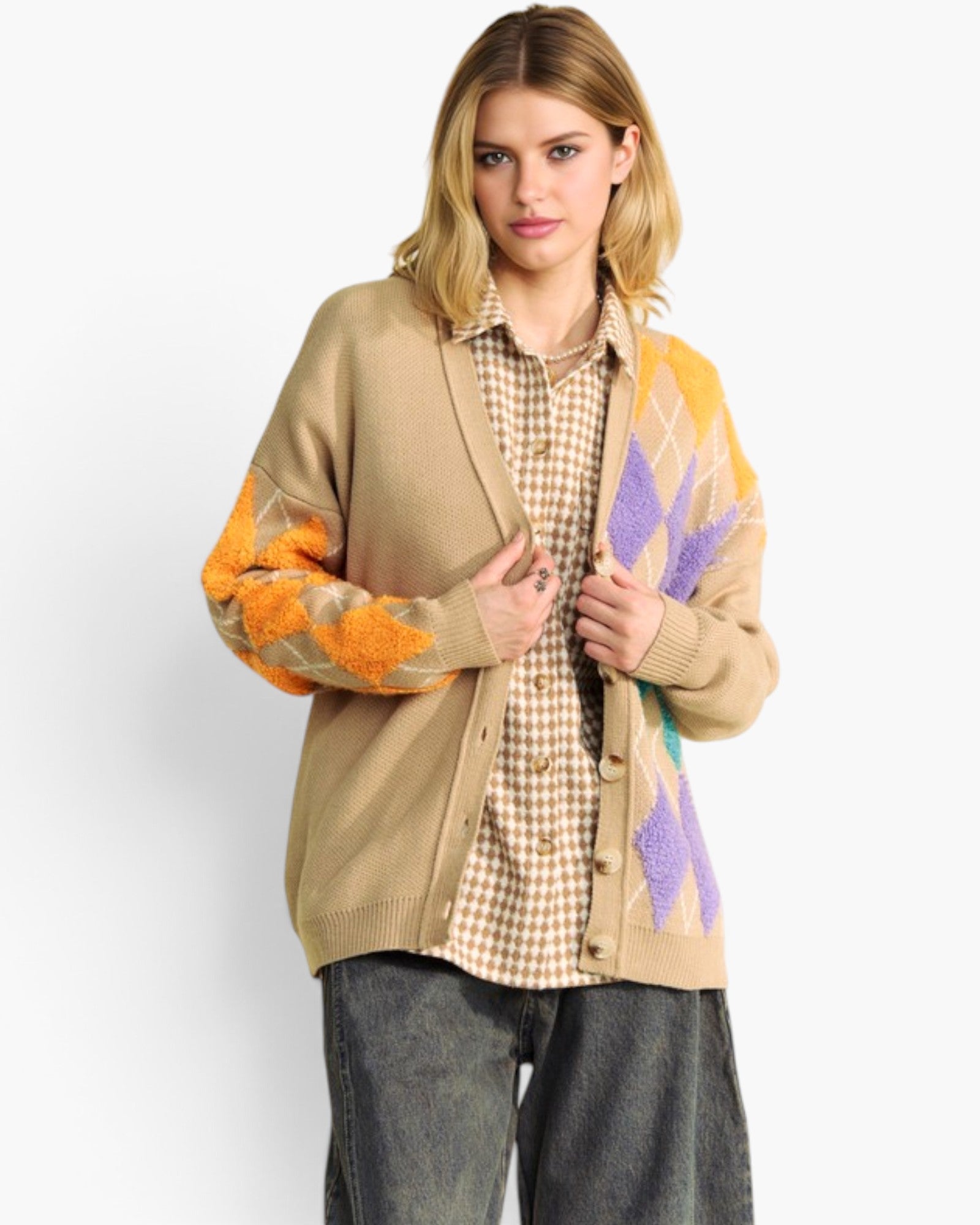 A woman models a beige cardigan with orange and purple argyle knit patterns on the sleeves and front panel, worn over a checkered shirt and dark gray jeans against a light gray studio background.