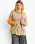A woman models a beige cardigan with orange and purple argyle knit patterns on the sleeves and front panel, worn over a checkered shirt and dark gray jeans against a light gray studio background.
