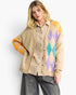 A woman models a beige knit cardigan with colorful argyle diamond patterns on the shoulders and sleeves, worn over a beige and white checkered shirt and gray pants against a light gray background.