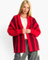 A blonde woman wearing a red and dark red striped open-front knit cardigan over a white top and gray jeans, photographed against a plain light gray studio background.