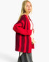 A blonde woman modeling a chunky knit cardigan with bold vertical stripes in bright red and deep burgundy, paired with a cream top and gray jeans against a plain white background.