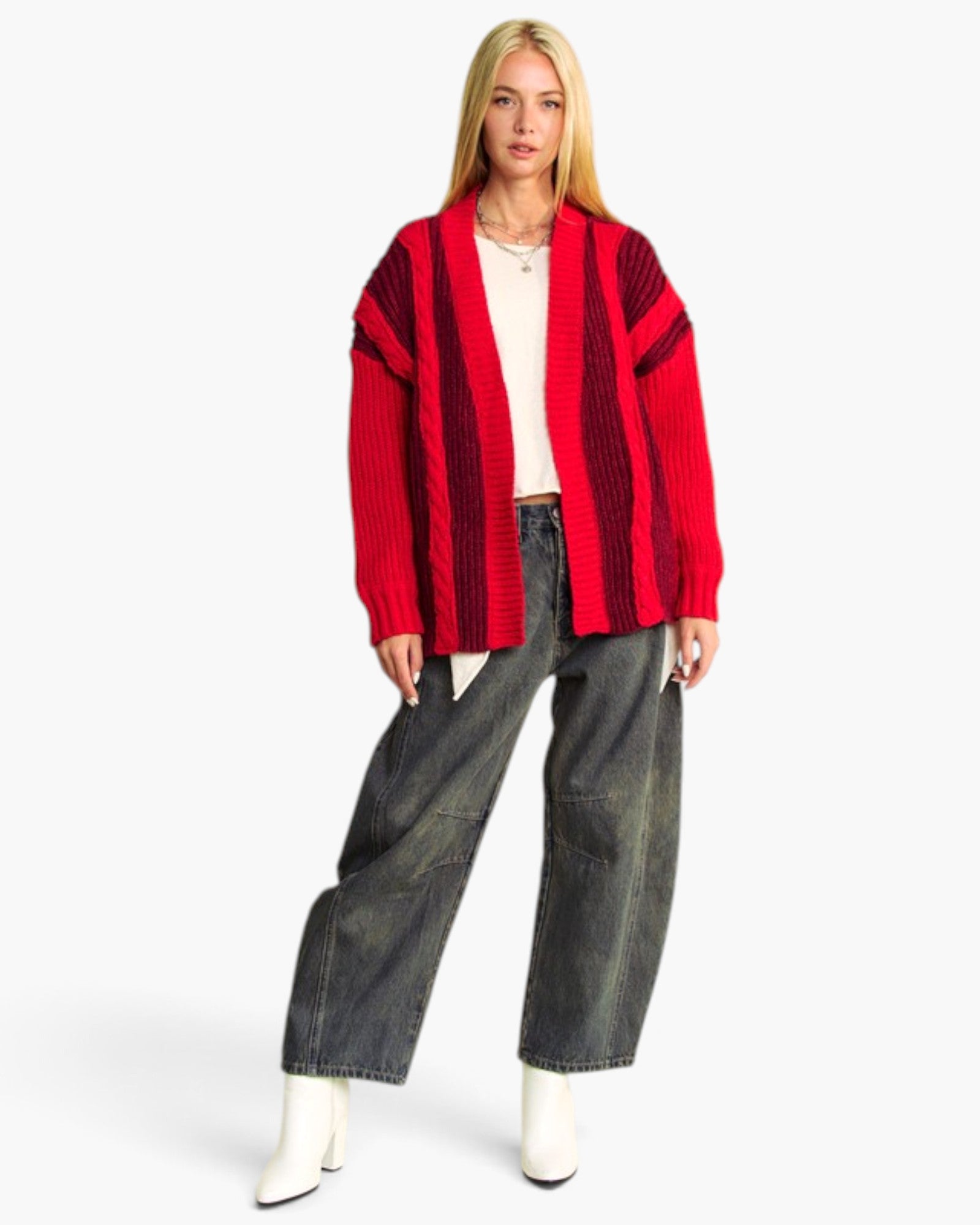 A blonde model wearing a red and black vertically striped open-front cardigan, a cream top, wide-leg dark gray jeans, and white ankle boots, standing against a light gray studio background.
