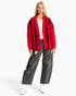 A blonde model wearing a red and black vertically striped open-front cardigan, a cream top, wide-leg dark gray jeans, and white ankle boots, standing against a light gray studio background.