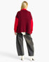 A woman shown from the back wearing a red and burgundy ribbed knit cardigan with wide-leg gray jeans and white heeled boots, against a plain light gray studio background.