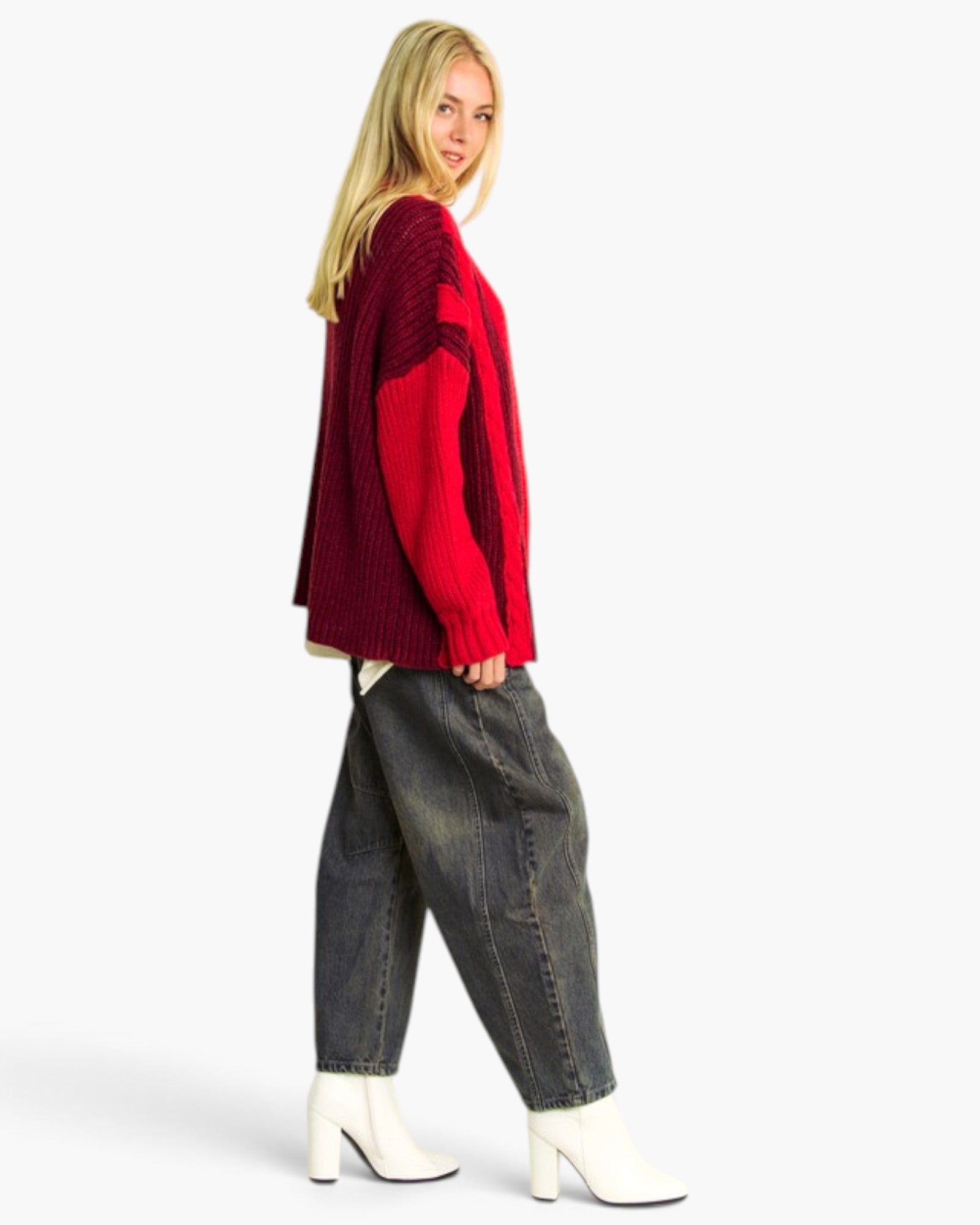 A woman modeling a red and burgundy color-blocked knit sweater, paired with dark gray baggy jeans and white heeled ankle boots, against a plain white studio background.