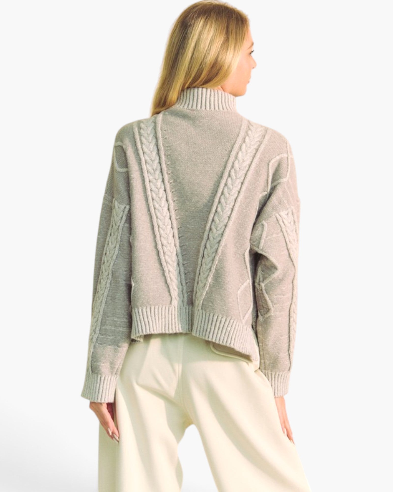 A woman shown from the back wearing a cropped, light taupe knit sweater with textured cable and diamond patterns, paired with cream-colored trousers, against a plain white background.