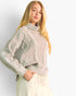 A blonde woman wearing a light beige cable-knit mock-neck sweater and cream drawstring trousers, photographed against a clean white studio background.