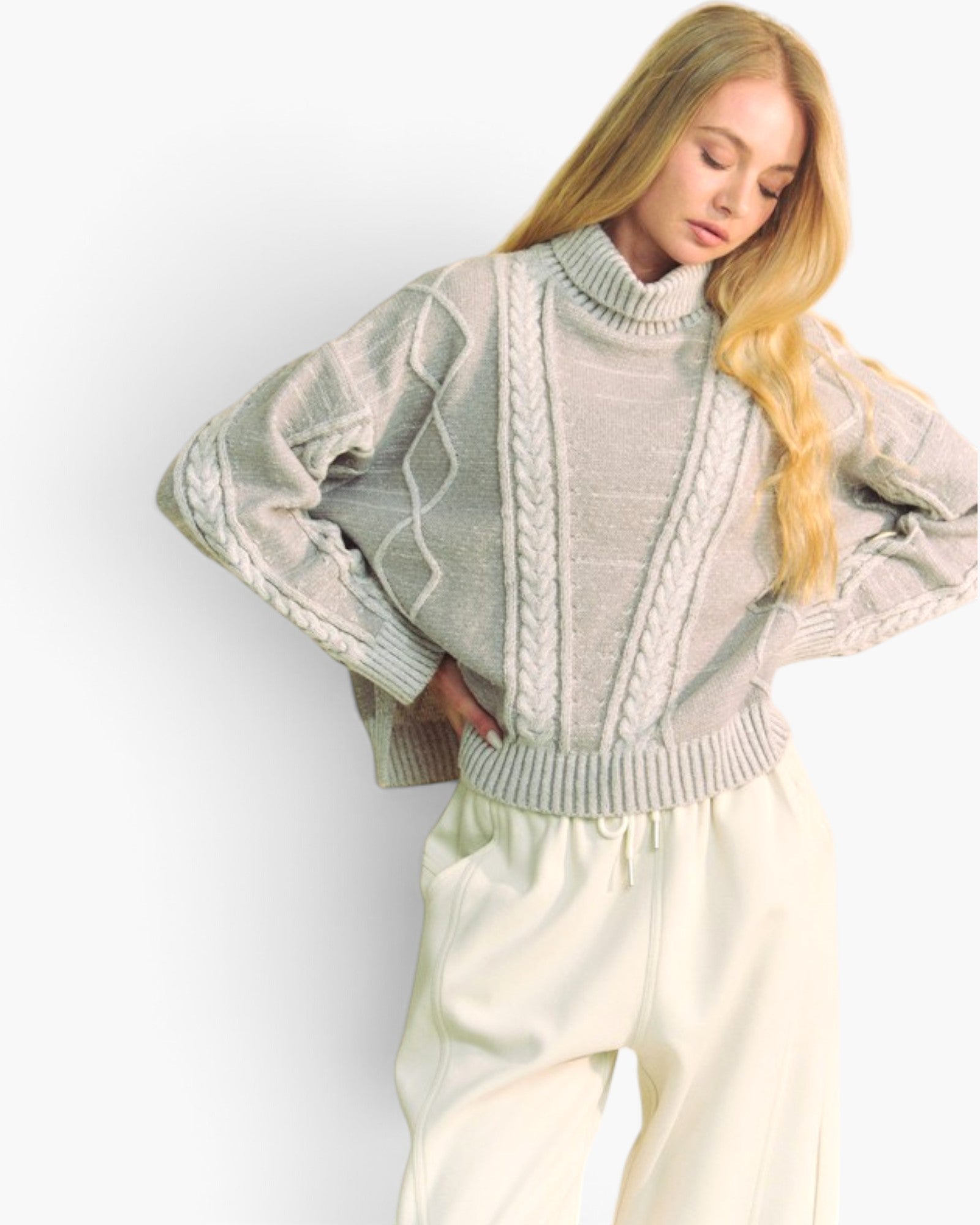 A woman wearing a light sage green cable-knit mock-neck sweater and cream-colored sweatpants, posing against a plain white studio background.