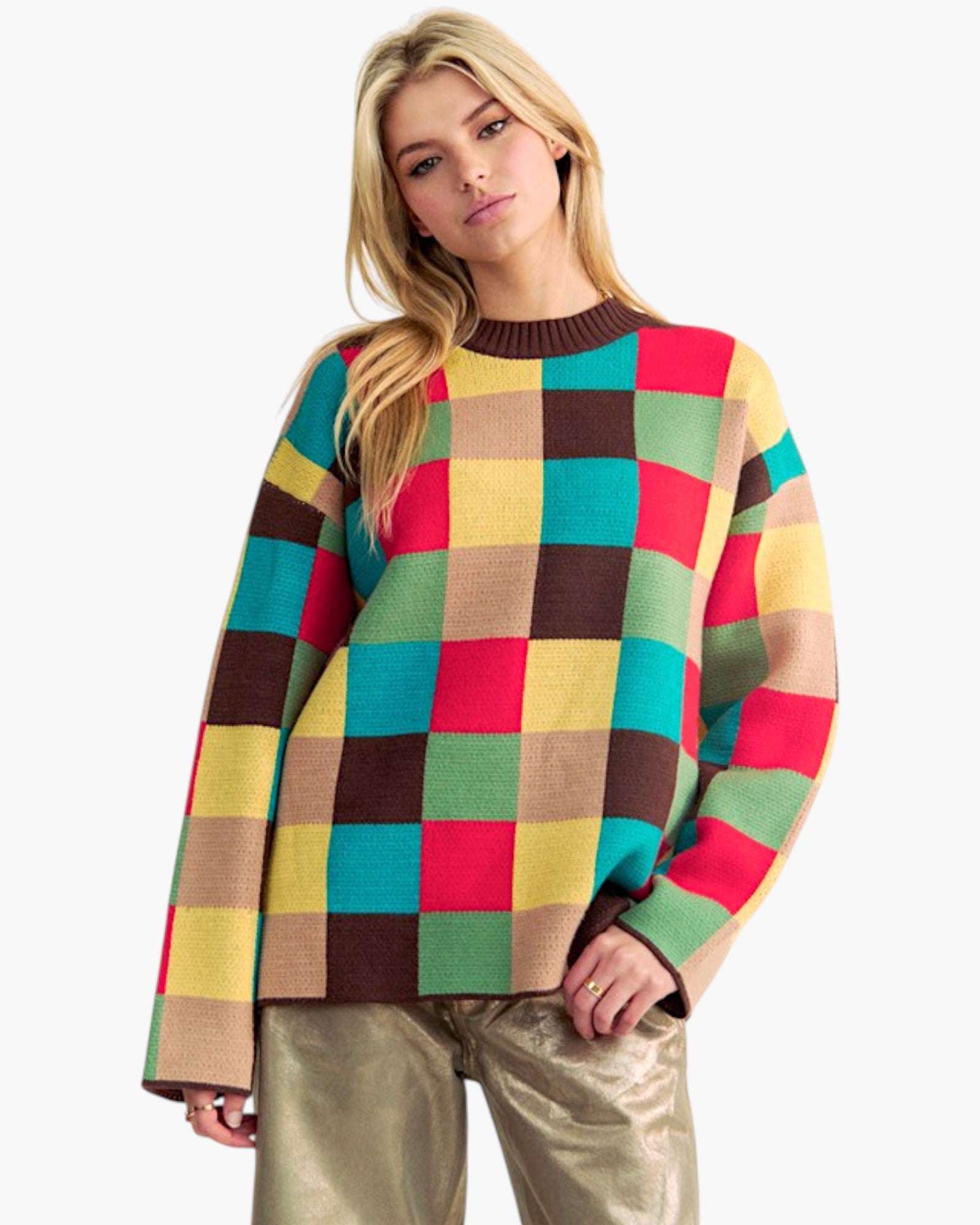 A woman wearing a colorful oversized knit sweater with a large checkered pattern in red, yellow, green, brown, and beige, paired with metallic gold pants, against a plain white background.