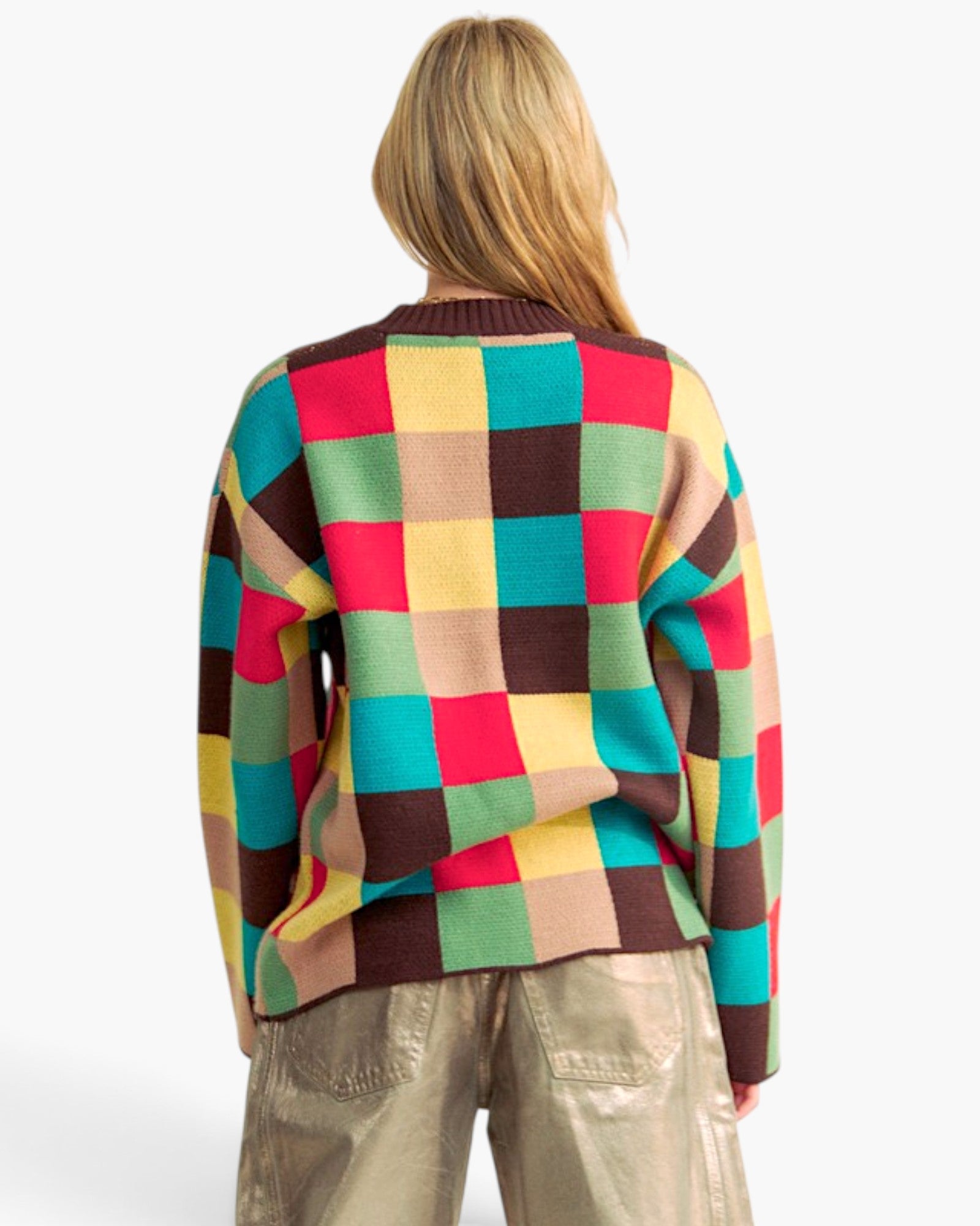 A woman viewed from behind wearing a colorful knit sweater with a large checkered pattern in yellow, green, teal, brown, and pink, paired with metallic beige pants against a plain white background.