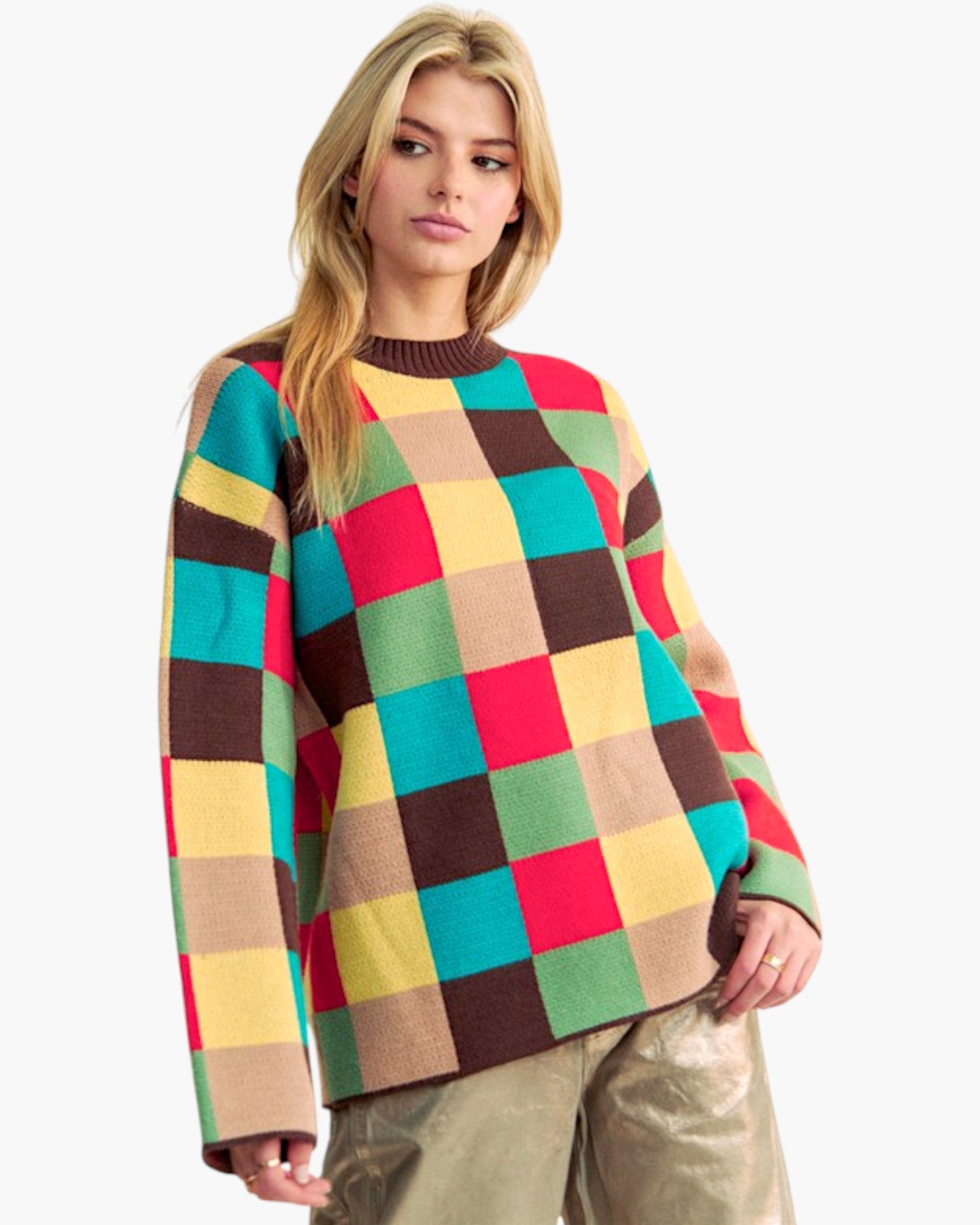 A woman wearing a colorful patchwork knit sweater with a large checkered pattern in red, yellow, green, brown, and beige, paired with metallic gold pants, against a plain white background.