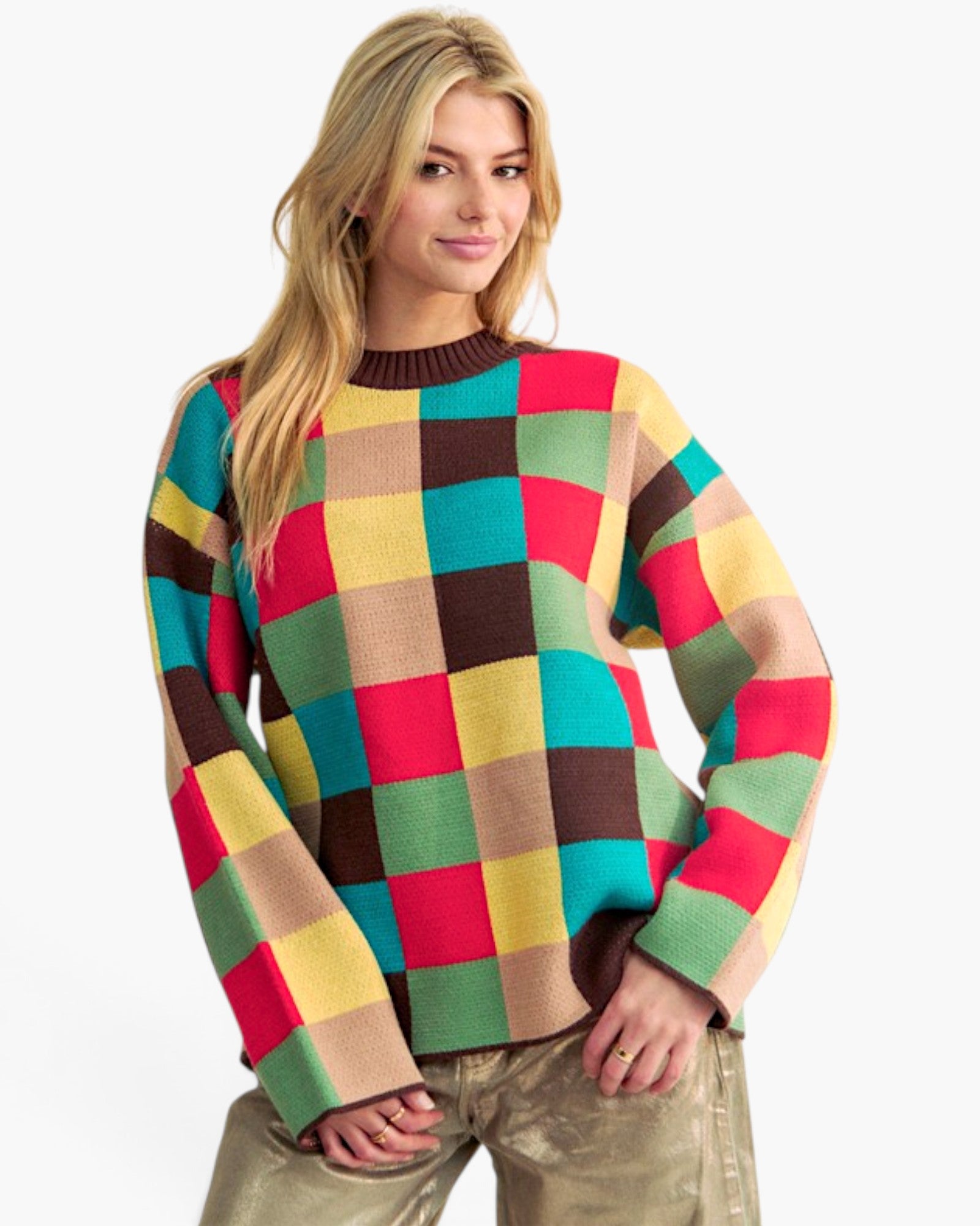 A woman wearing a colorful long-sleeve knit sweater with a large checkered pattern in red, green, yellow, beige, and brown, paired with metallic gold pants, against a plain white background.
