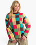 A woman wearing a colorful long-sleeve knit sweater with a large checkered pattern in red, green, yellow, beige, and brown, paired with metallic gold pants, against a plain white background.