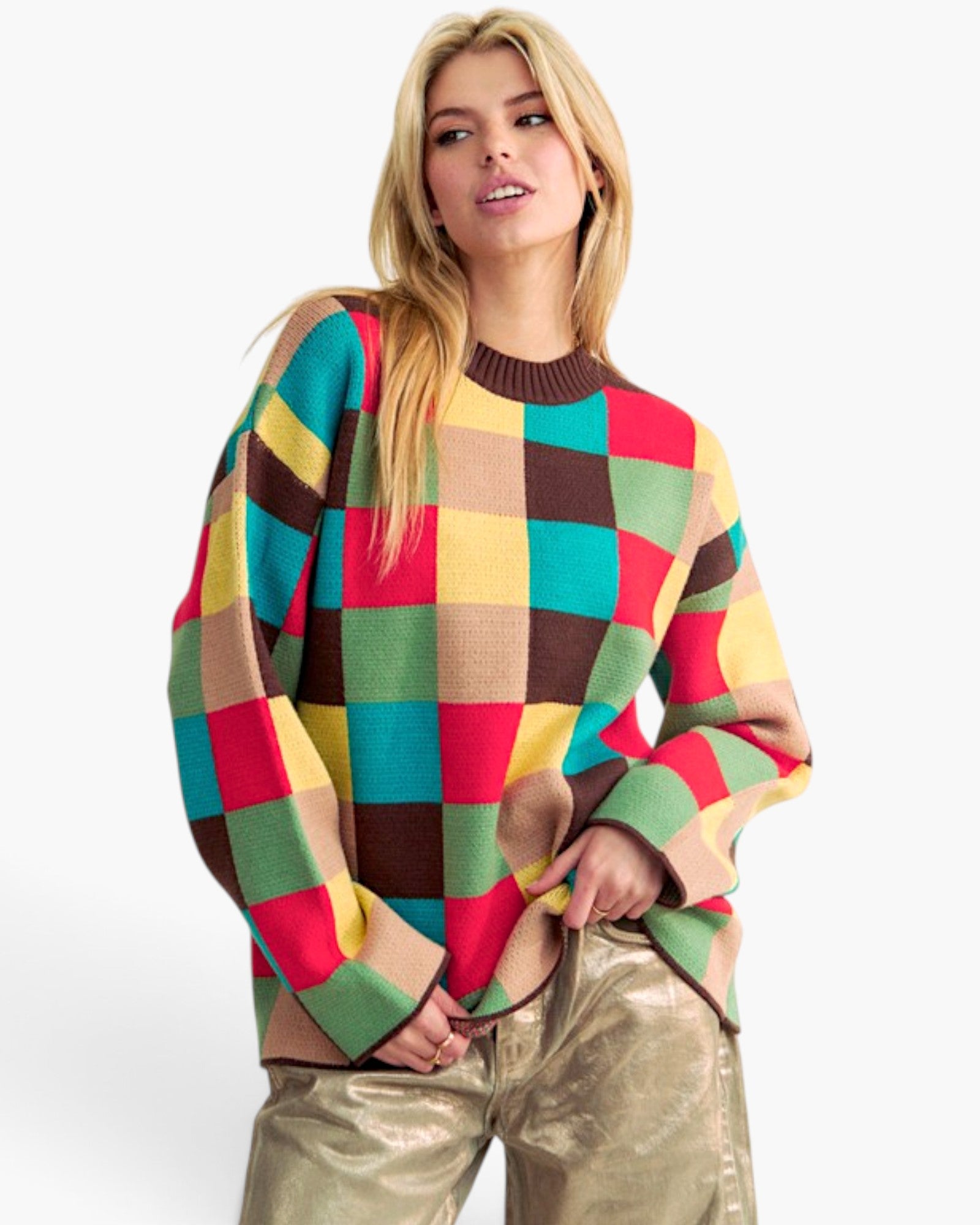 A woman modeling a colorful oversized knit sweater with a large checkered pattern in red, green, yellow, brown, and beige, paired with metallic gold pants against a plain white background.